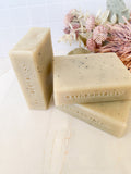 Three bars of Bath Body Bliss stamped soap on a light surface with decorative flowers in the background.