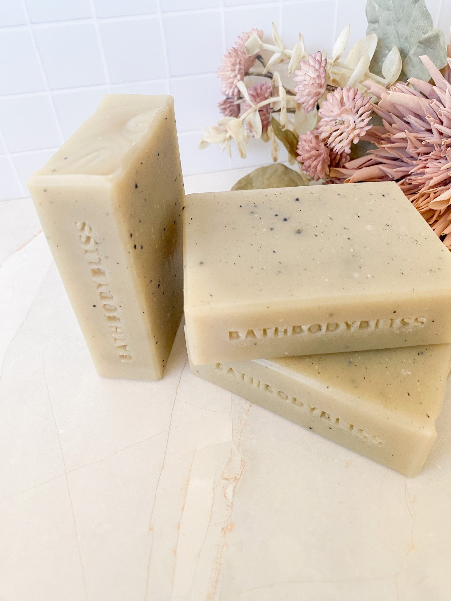 Three bars of Bath Body Bliss stamped soap on a light surface with decorative flowers in the background.