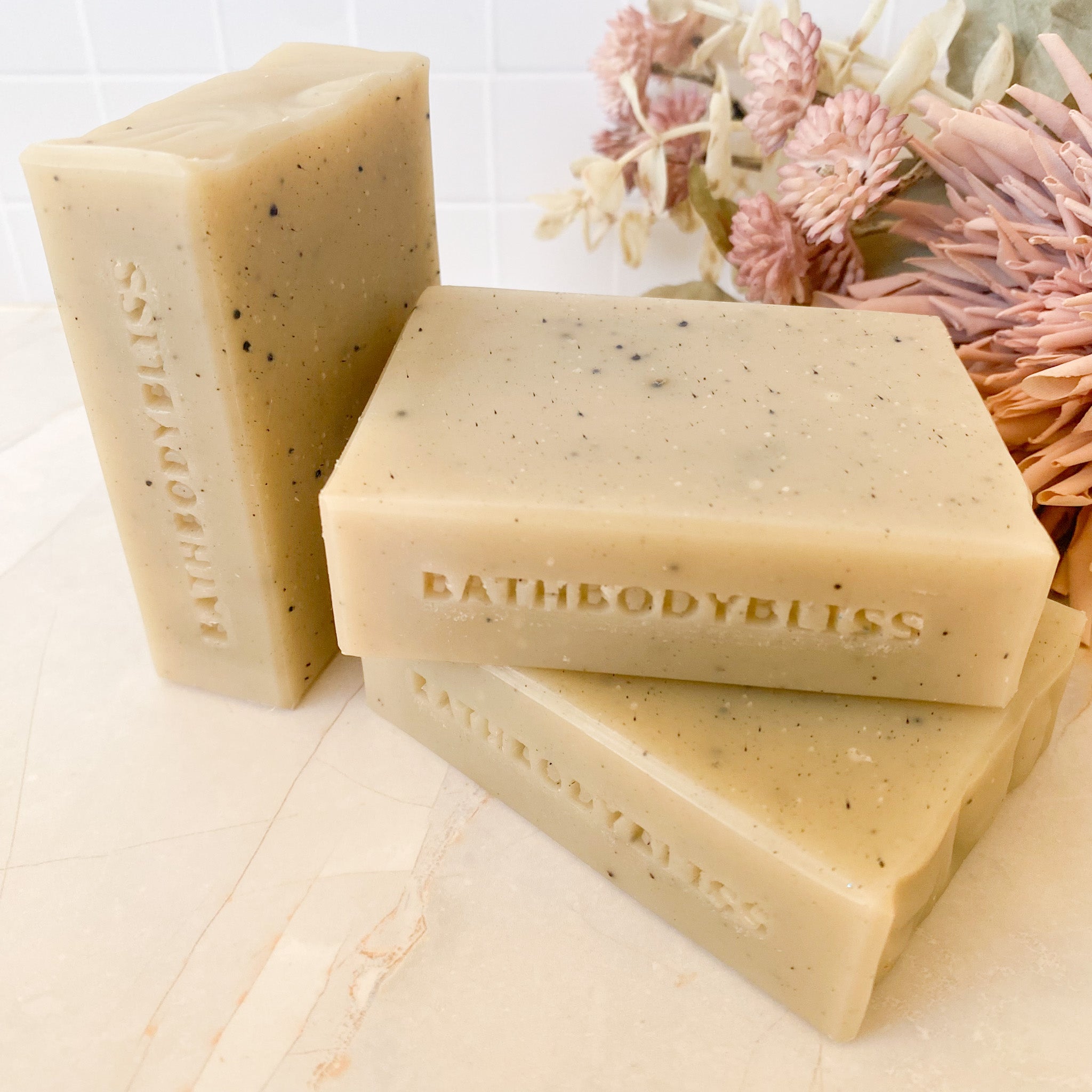 Three bars of BathBodyBliss soap on a wooden surface with decorative flowers in the background.