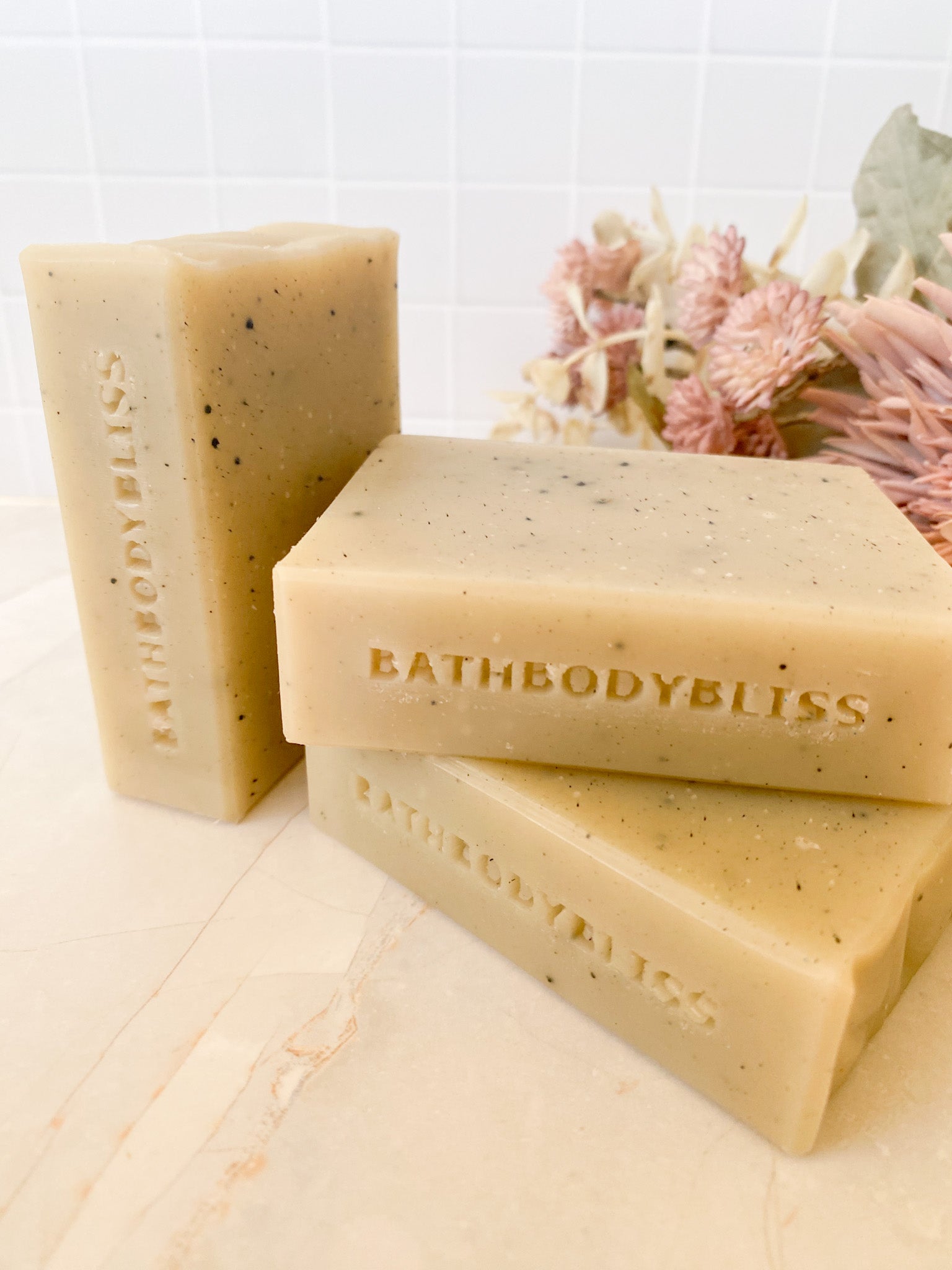 Three bars of BathBodyBliss soap stacked on a marble surface with a white tiled background.