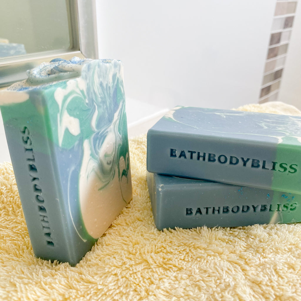 three bars of bathbody bliss stamped soaps in blue, green and white swirls, on a yellow towel with bathroom tiles in the background