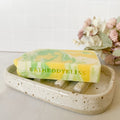 Marbled yellow and green soap bar on a speckled ceramic soap dish with 'BATHBODYBLISS' branding, against a light background.