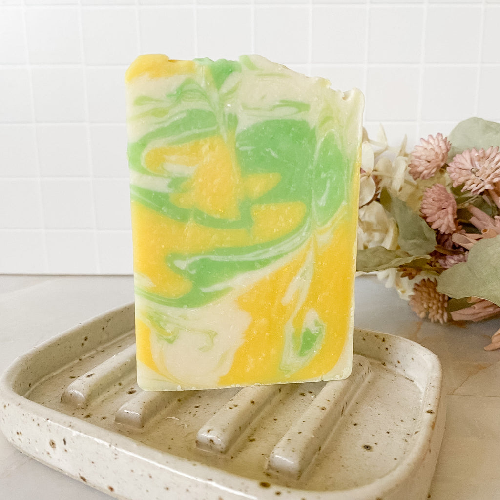 Swirled yellow and green soap bar on a textured soap dish with flowers in the background.