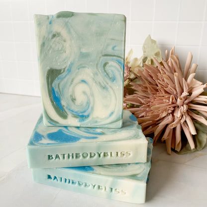 Stack of blue, green and white swirld Bathbodybliss soap bars with a decorative flower on a white surface.