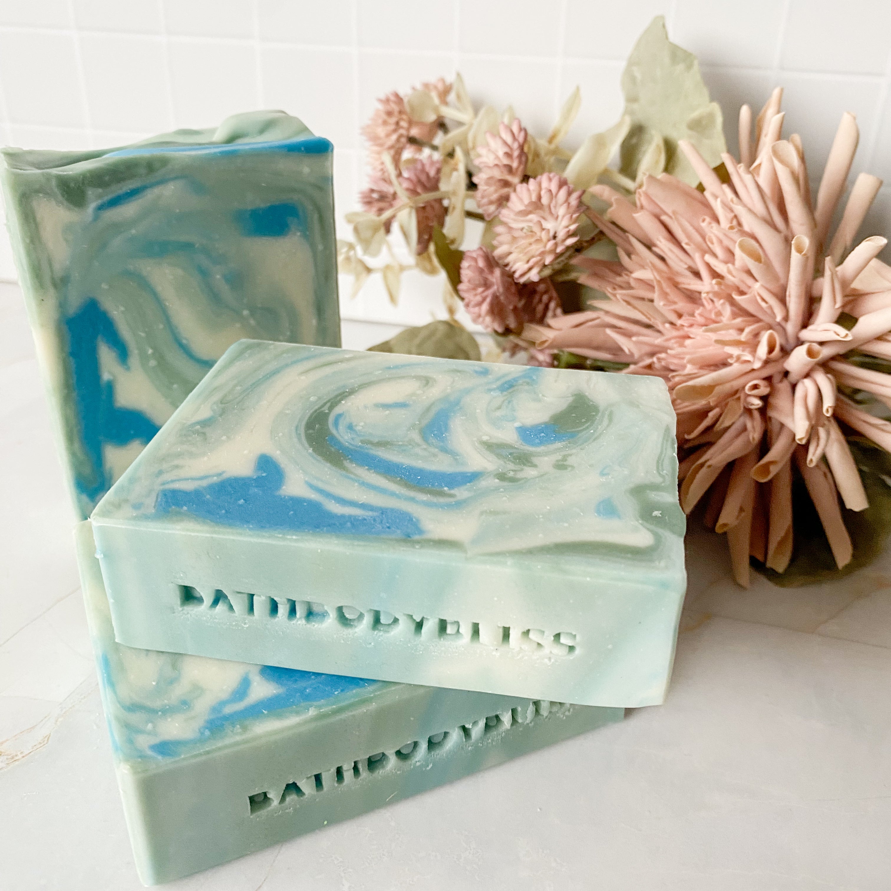 Three swirled soap bars with 'BathBodyBliss' branding on a white surface with flowers in the background.