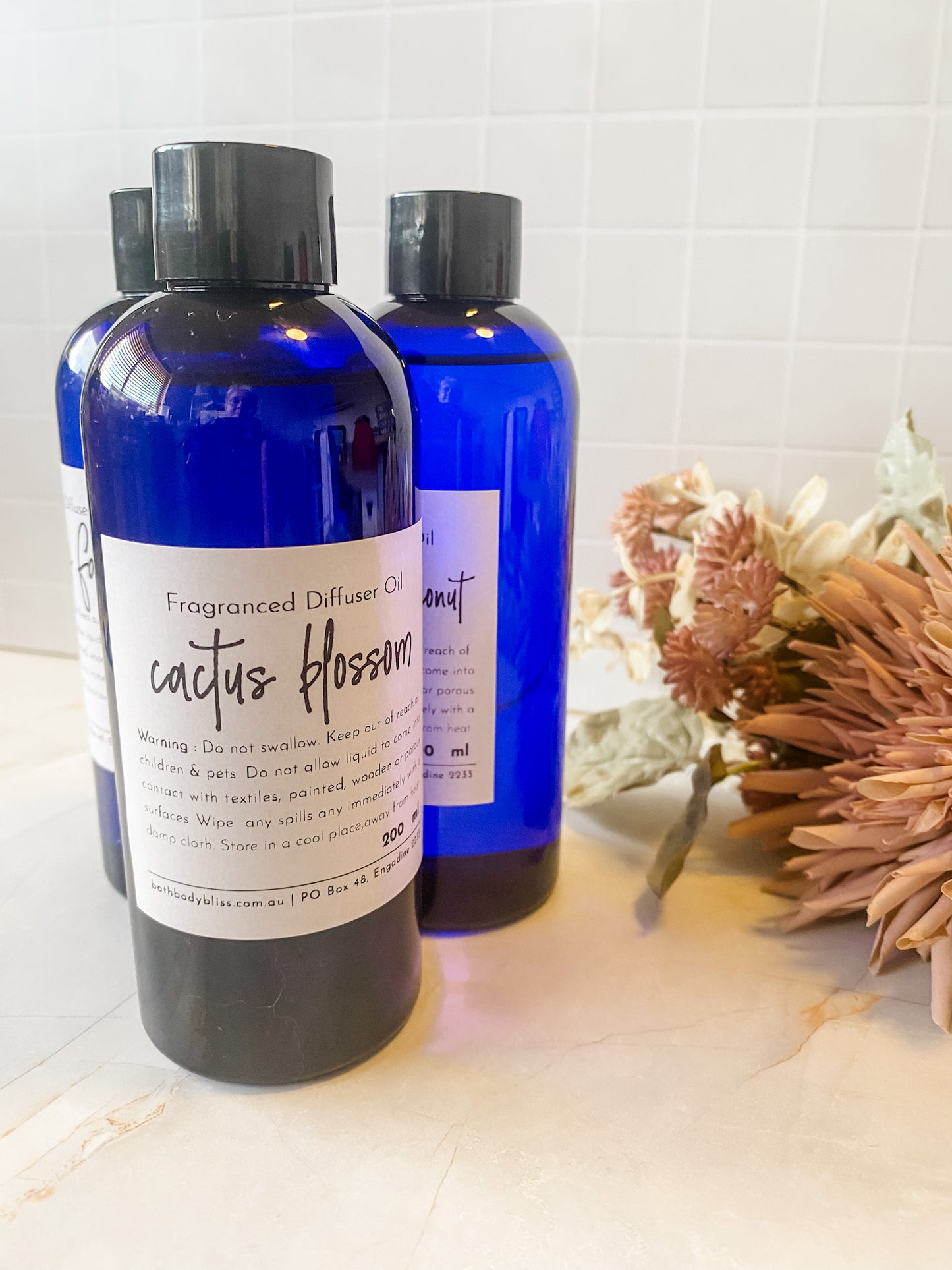 Three blue bottles of diffuser oil on a white surface with a decorative plant in the background.