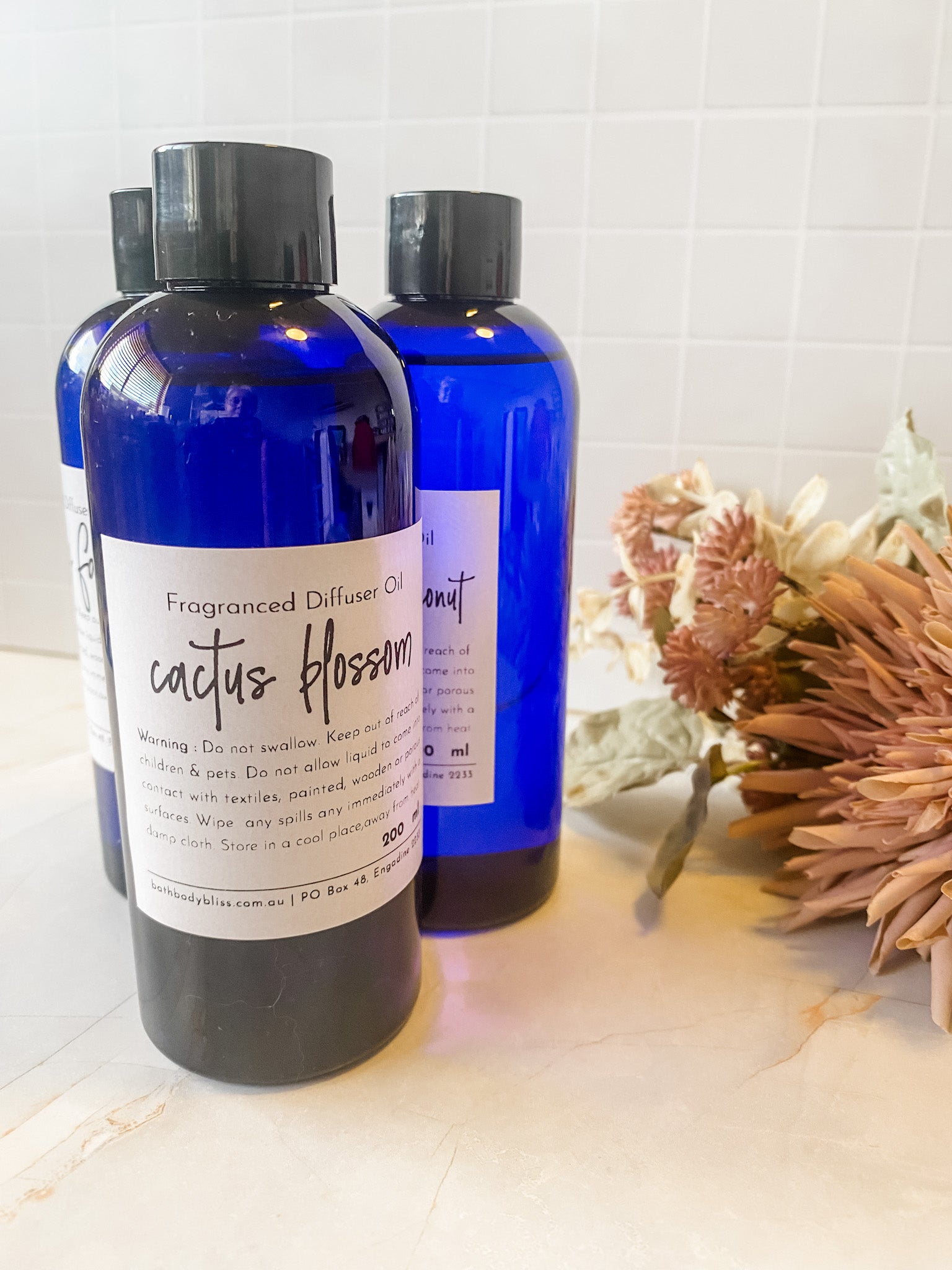 Three blue bottles of diffuser oil on a white surface with a decorative plant in the background.