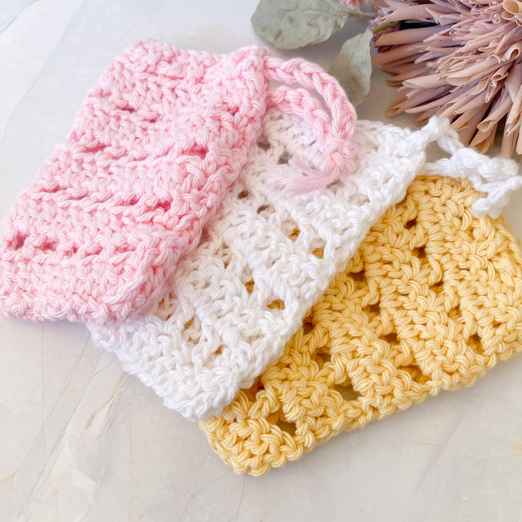 Three crocheted soap savers in pink, white, and yellow on a light marble surface.