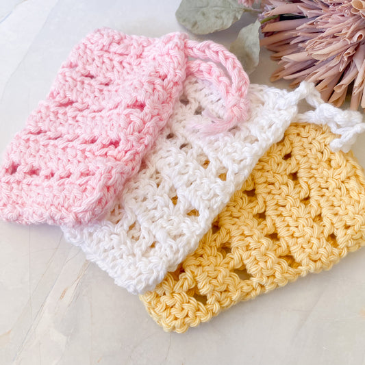 Three crocheted soap savers in pink, white, and yellow on a light marble surface.