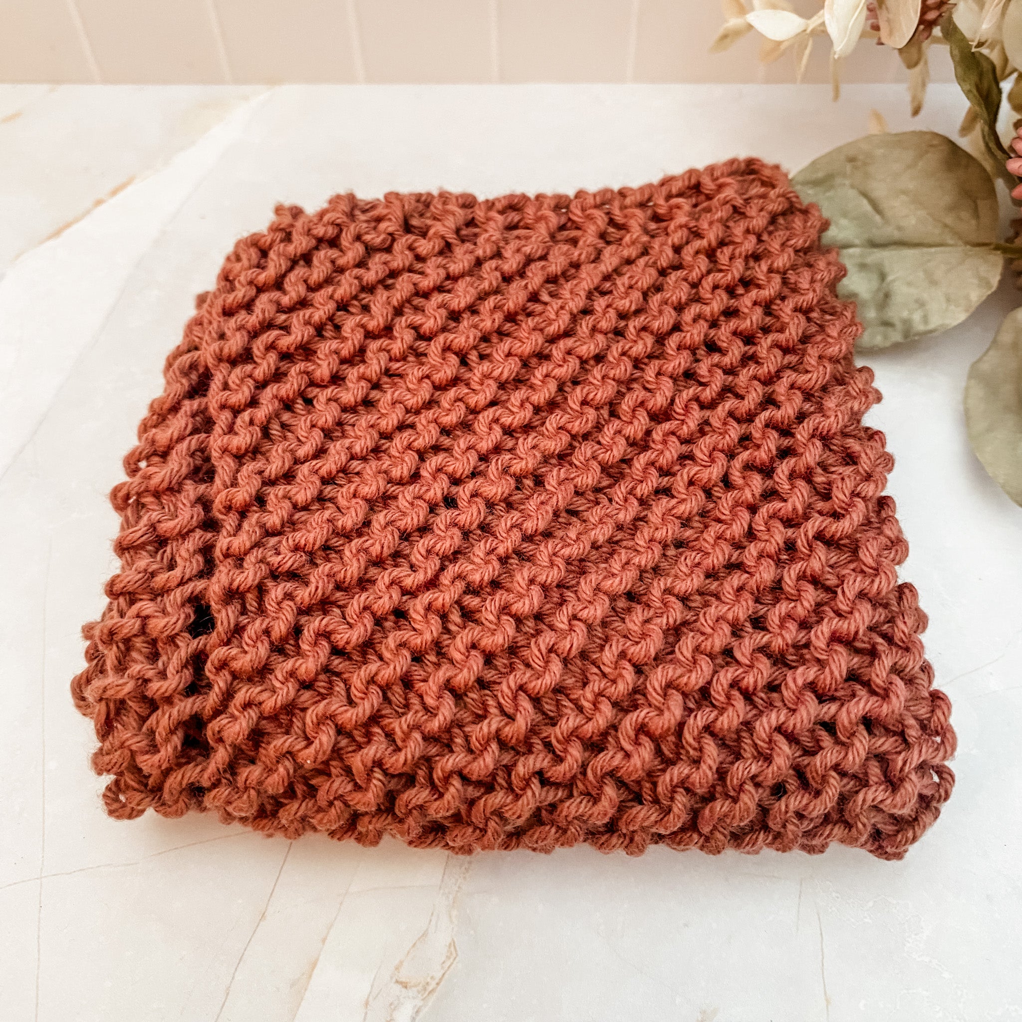 A hand-knitted brown cotton face or dish cloth laid out on a marble surface.