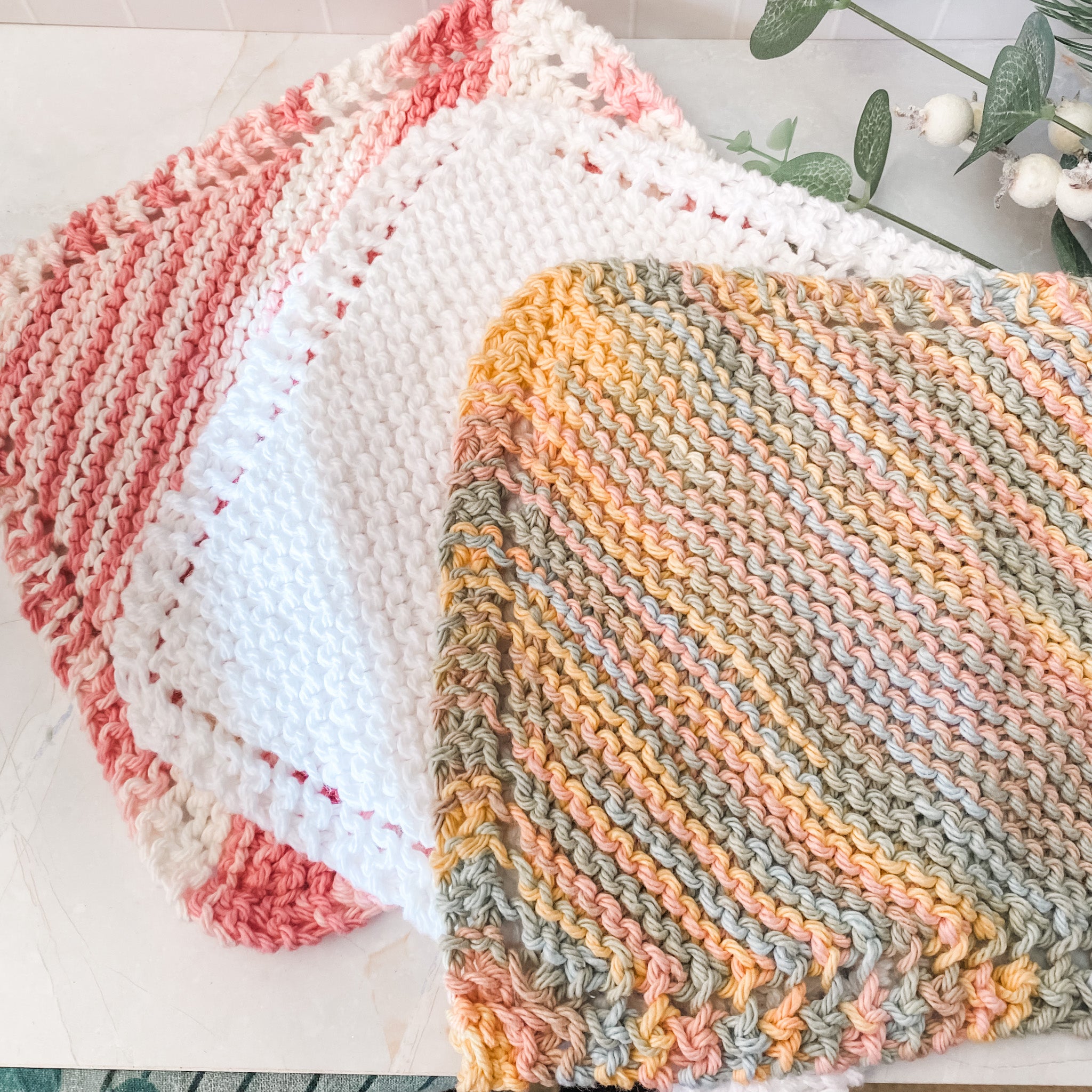 Three knitted dishcloths in different colors on a light surface with greenery.