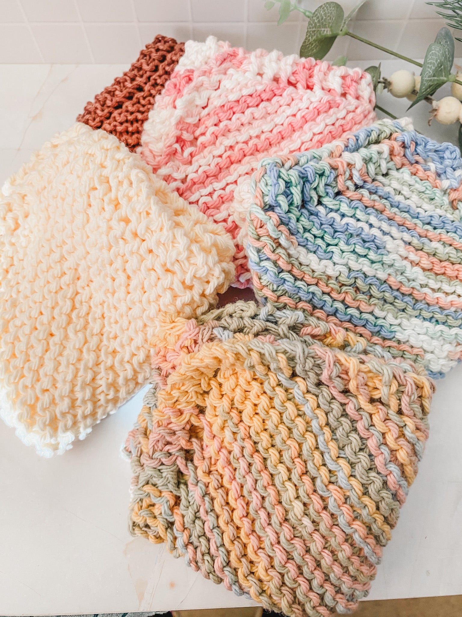 Stack of colorful knitted dishcloths on a white surface with a plant in the background.
