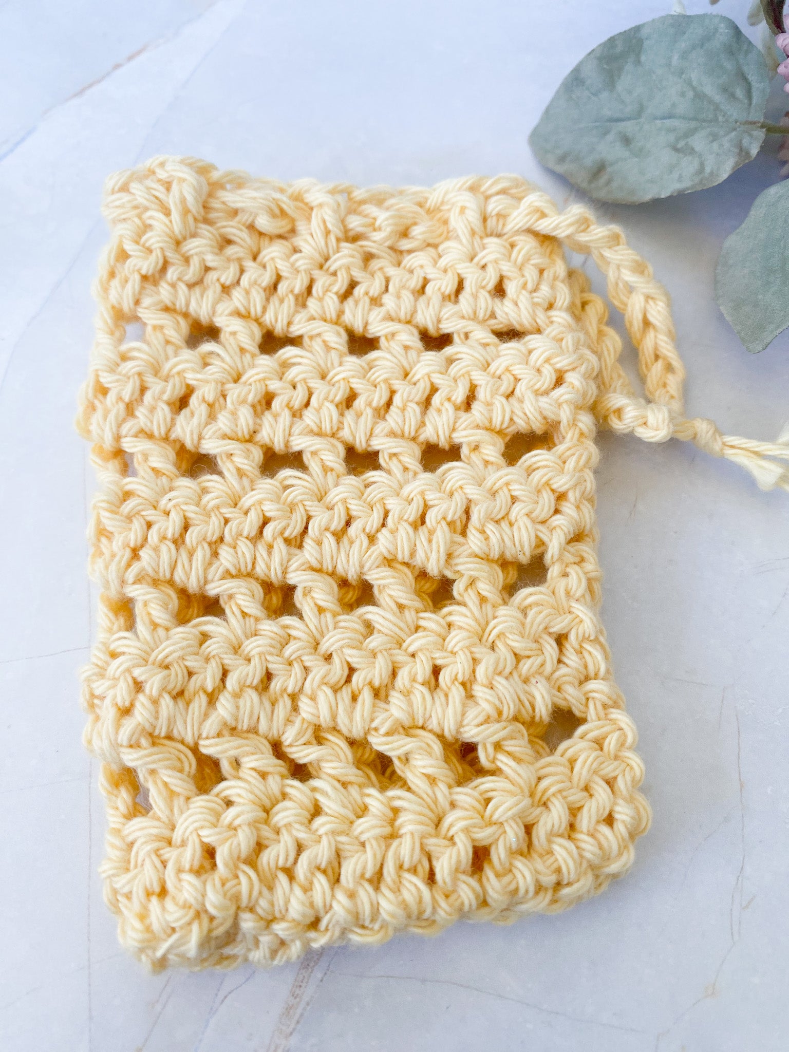 Yellow crocheted soap saver sack on a white surface with green leaves in the background