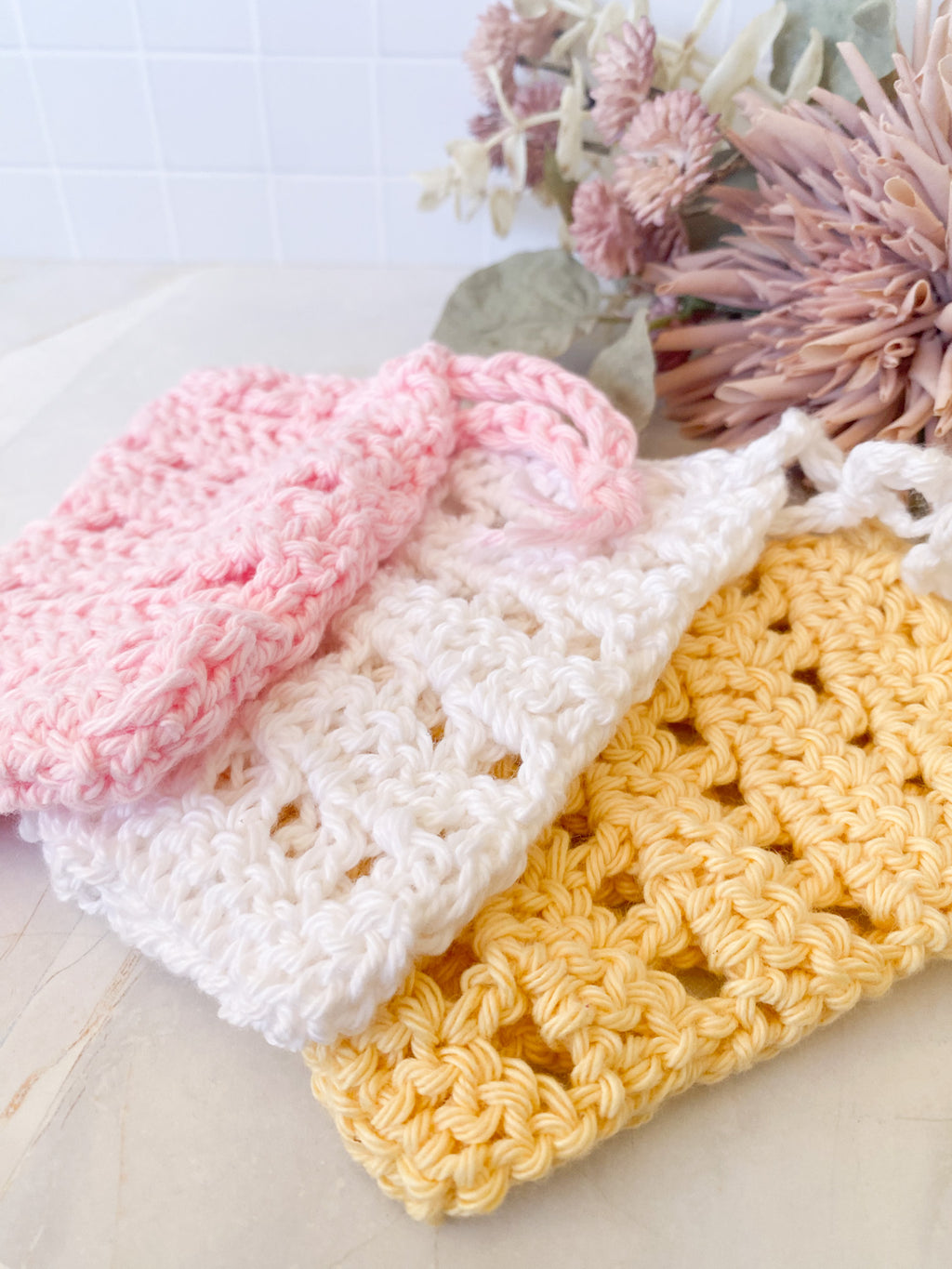 Three crocheted soap saver sacks in pink, white, and yellow on a light marble surface with flowers in the background.