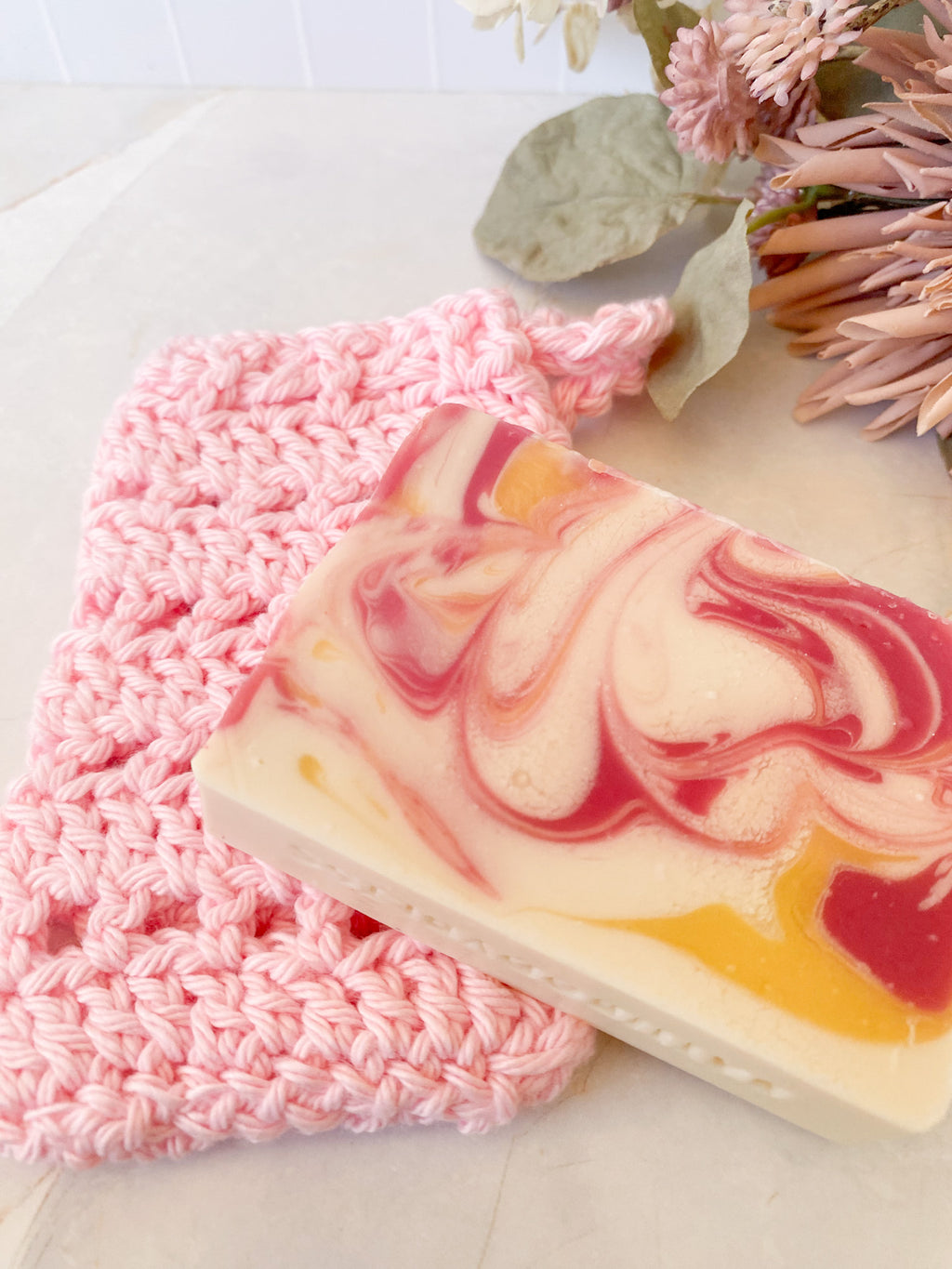 Marbled soap bar with pink and yellow swirls on a pink crochet soap saver sack, with dried flowers in the background.