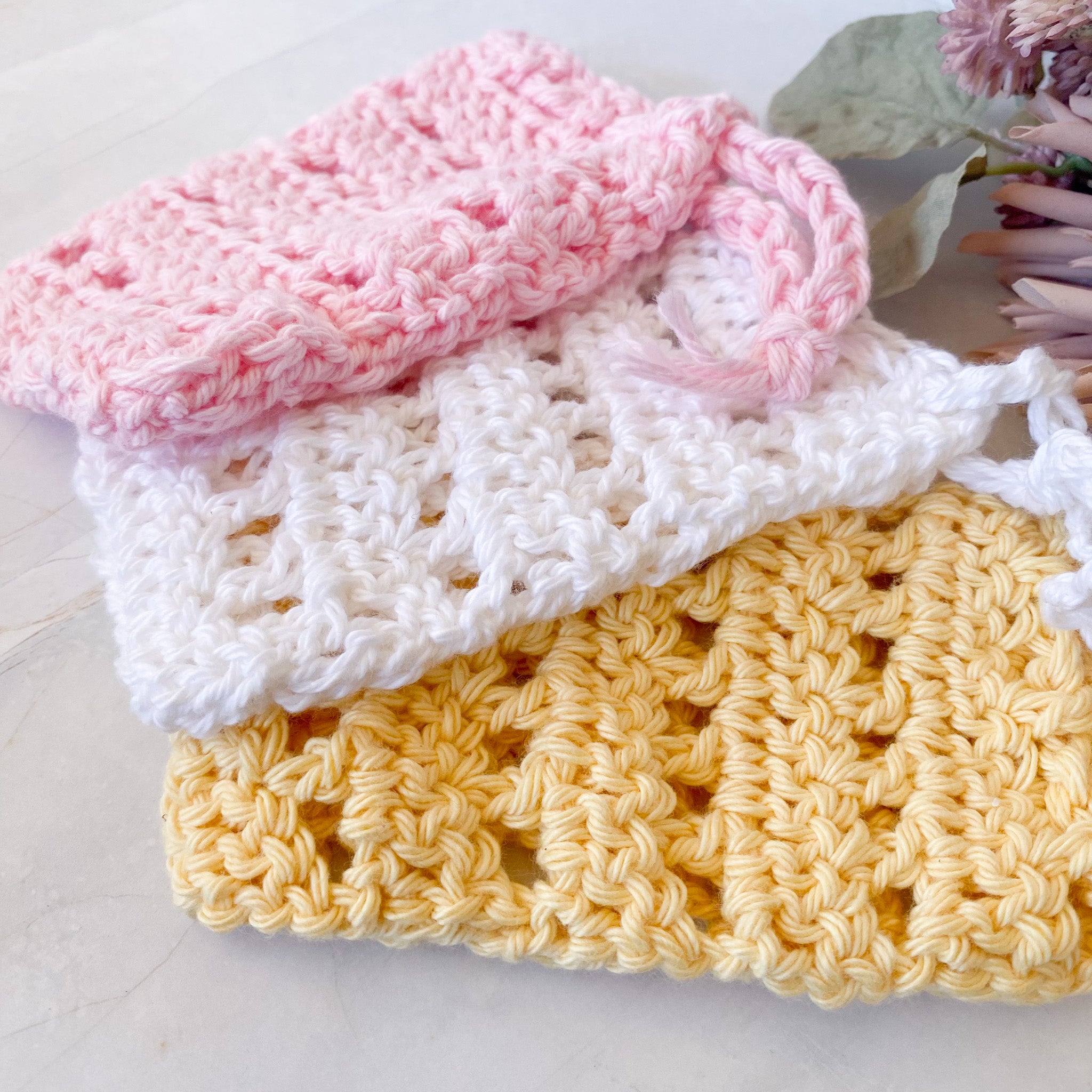 Stack of crocheted soap saver sacks in pink, white, and yellow on a light surface.