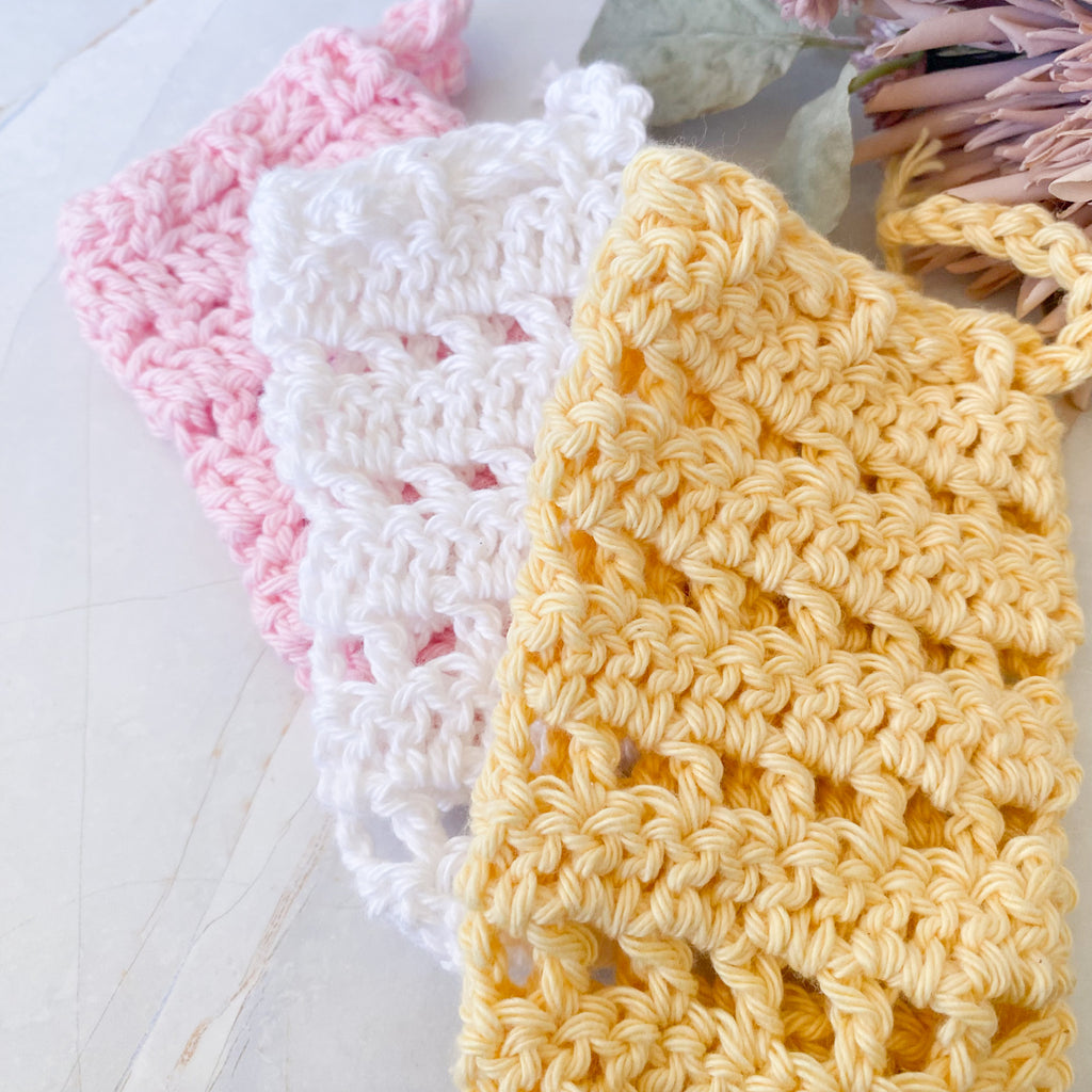 Three crocheted soap saver sacks in pink, white, and yellow on a light marble surface.