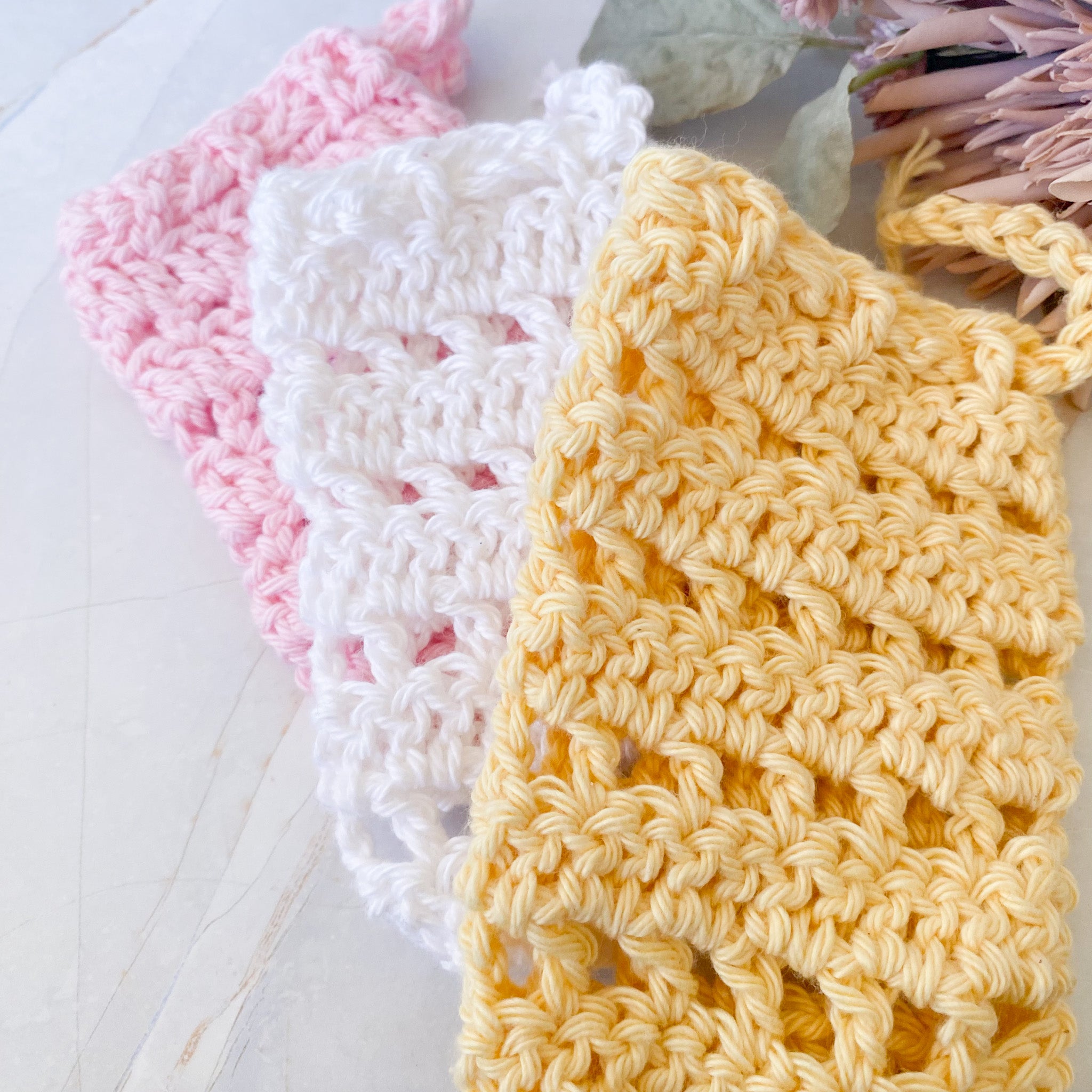 Three crocheted soap saver sacks in pink, white, and yellow on a light marble surface.