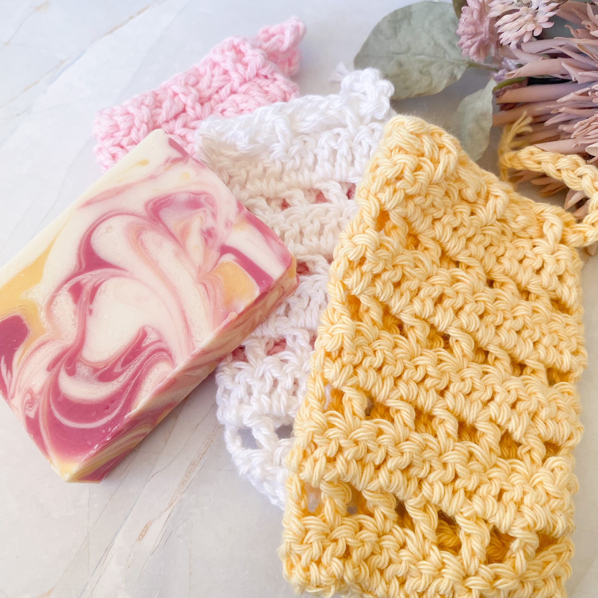 Bar of soap with pink and white swirls next to a yellow crocheted soap saver sack on a light wooden surface.