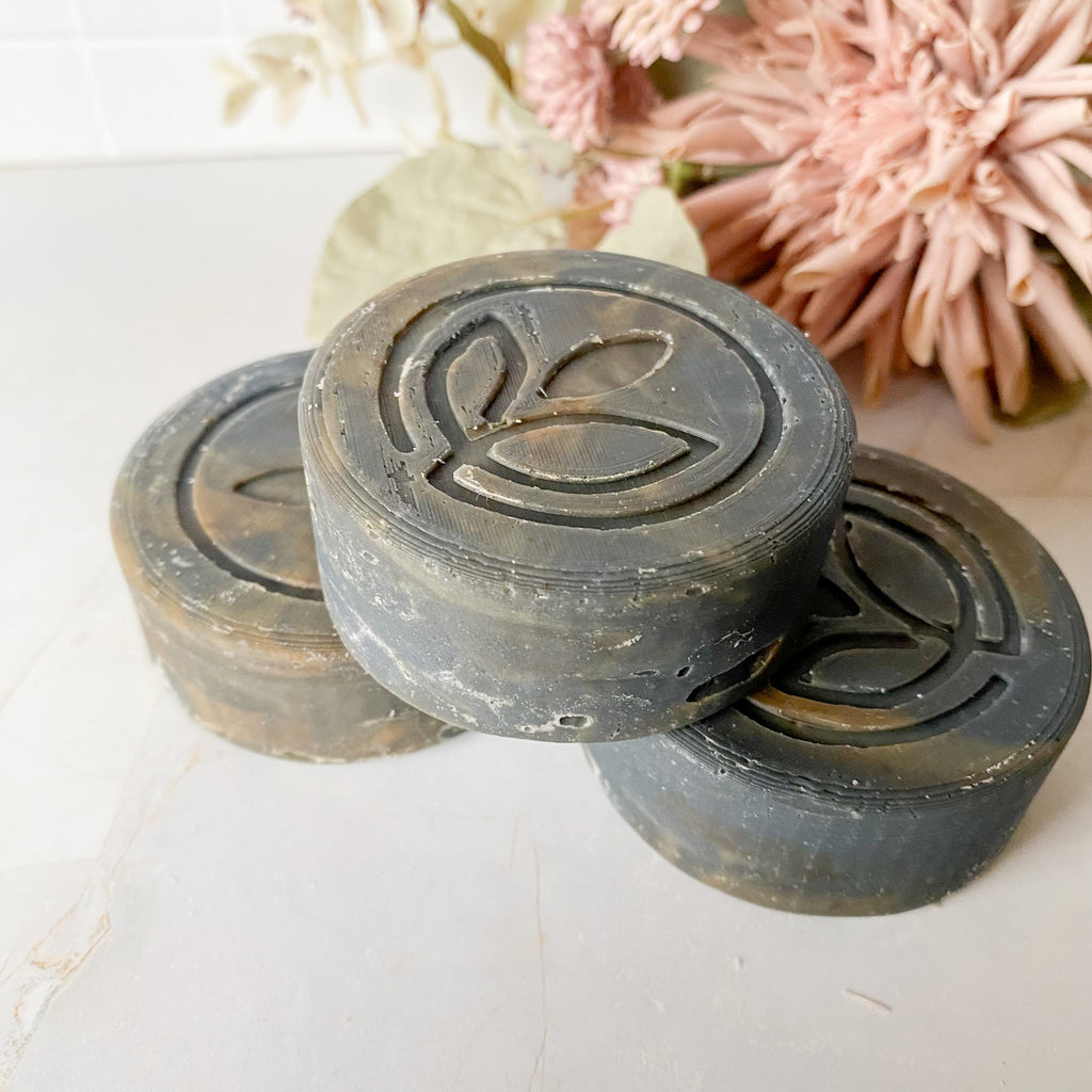 Three dark grey swirled soap bars with a leaf design on a light surface with pink flowers in the background