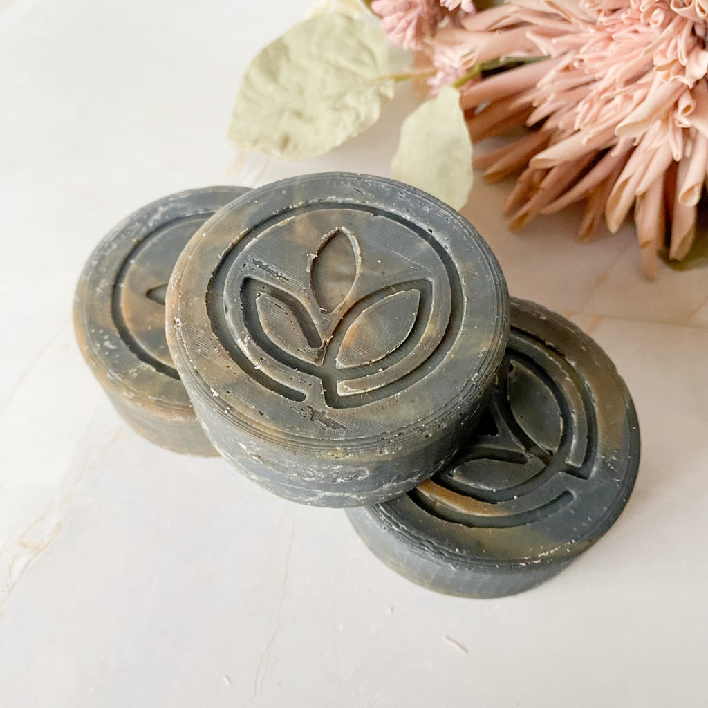 Three dark gray round soap bars with a logo on a light surface with flowers in the background
