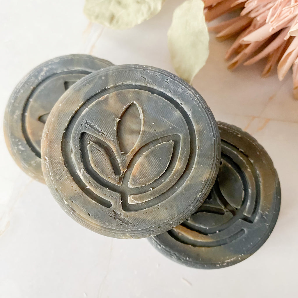 Three circular dark grey swirled soap bars with a leaf logo design on a light background