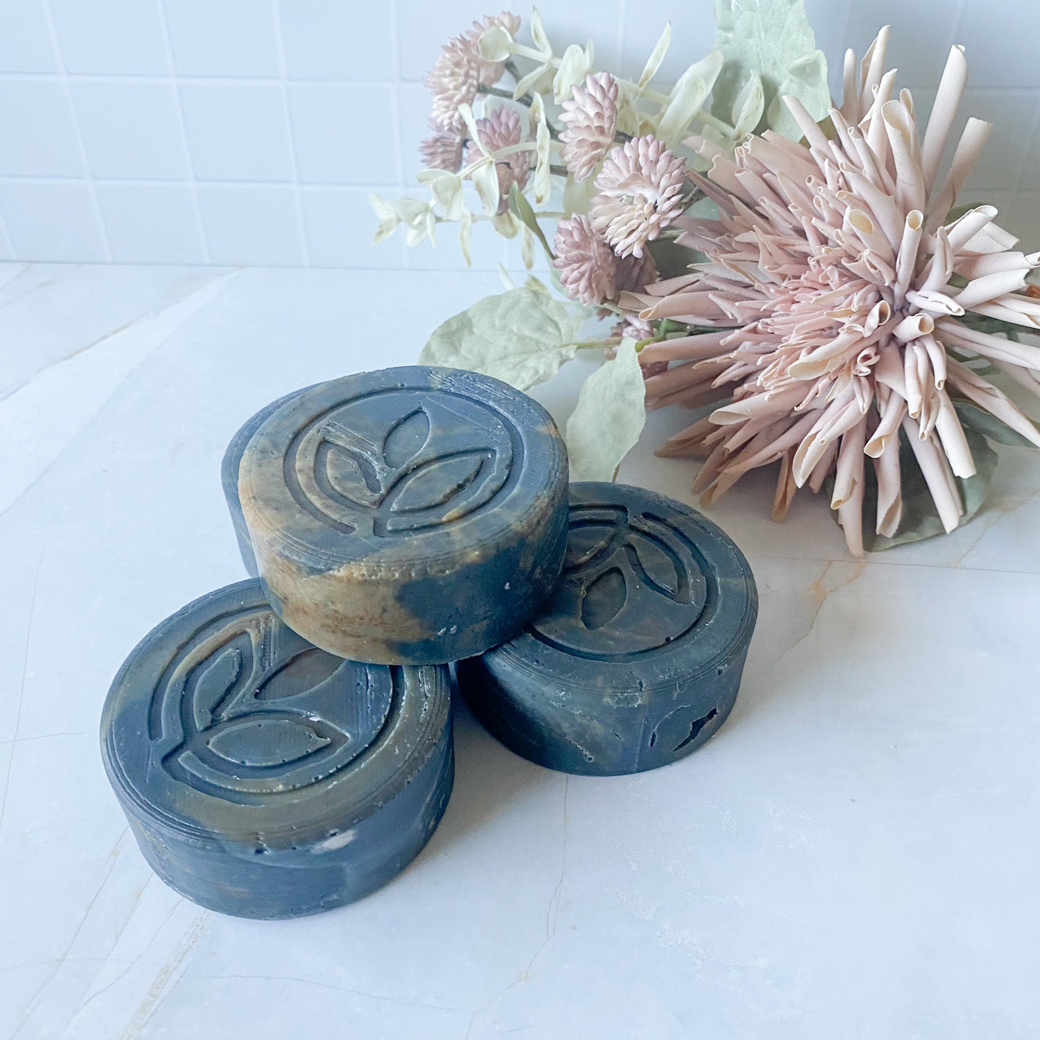 stack of four dark grey swirled round soap bars with leaf design on a white surface with flowers in the background