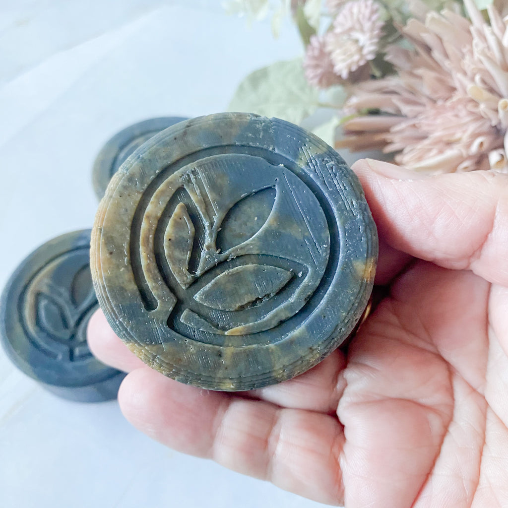 Hand holding a round bar of dark grey swirled soap with a leaf logo design, surrounded by flowers on a light background