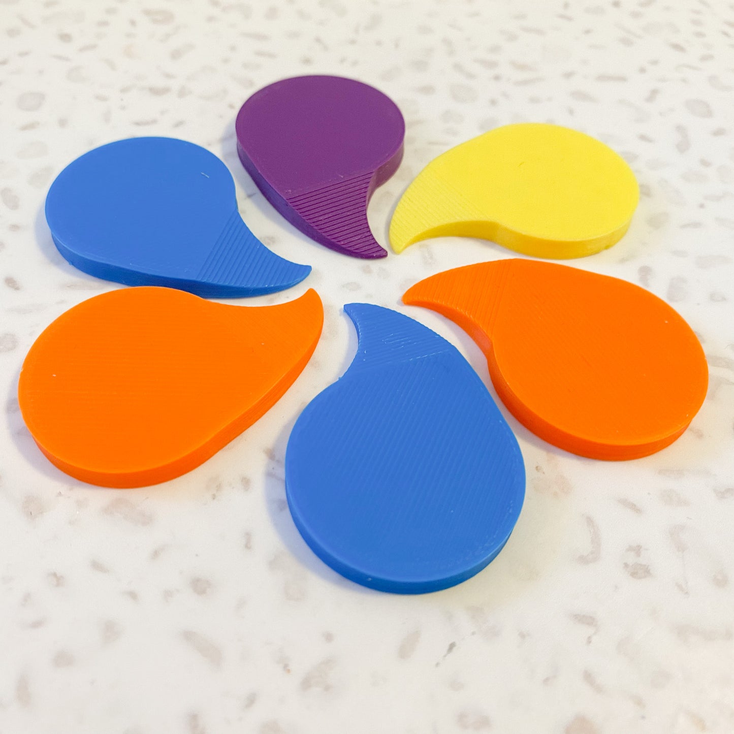 six brightly coloured 3D printed plastic shrink wrap openers arranged in a circle on a pale speckled background