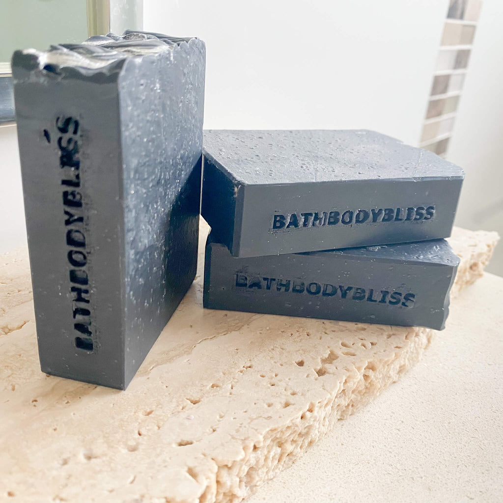 Three dark gray soap bars with 'BathBodyBliss' branding on a marble surface.