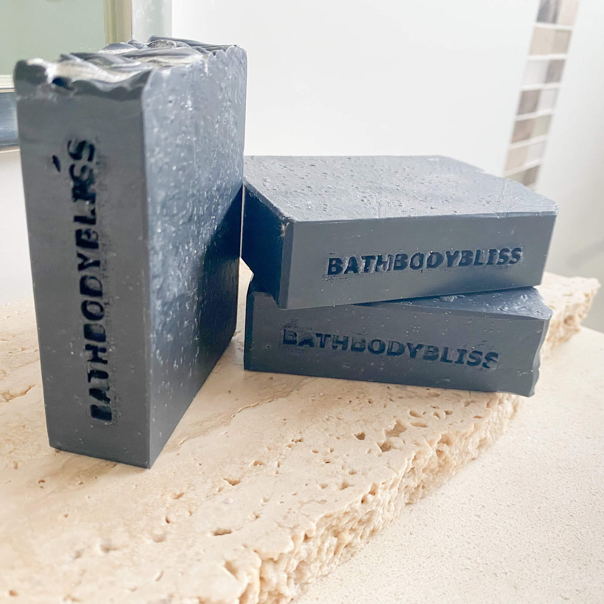 Three dark gray soap bars with 'BathBodyBliss' branding on a marble surface.