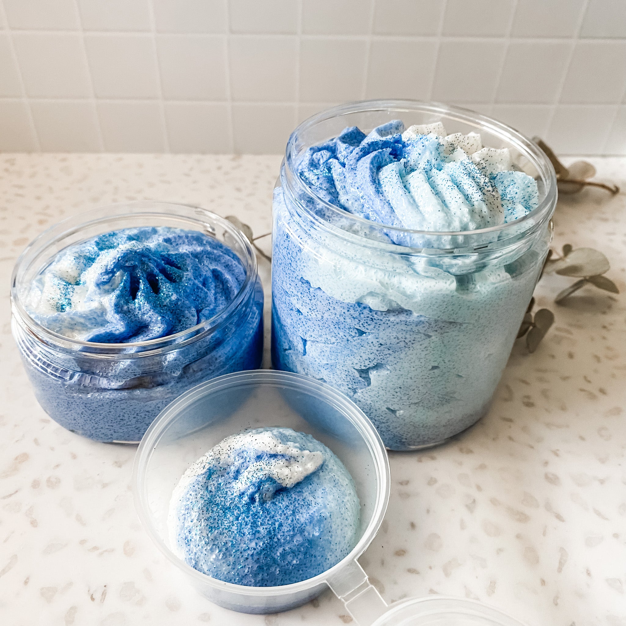 three different size containers of whipped body scrub in shades of blue, arranged on a speckled background
