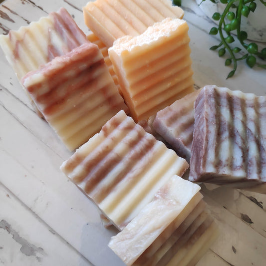 A variety of handcrafted guest soaps with organic shea and cocoa butter, displayed on a wooden surface. The soaps have a marble-like appearance with different colors including white, pink, purple, and orange, and are adorned with textured ridges.