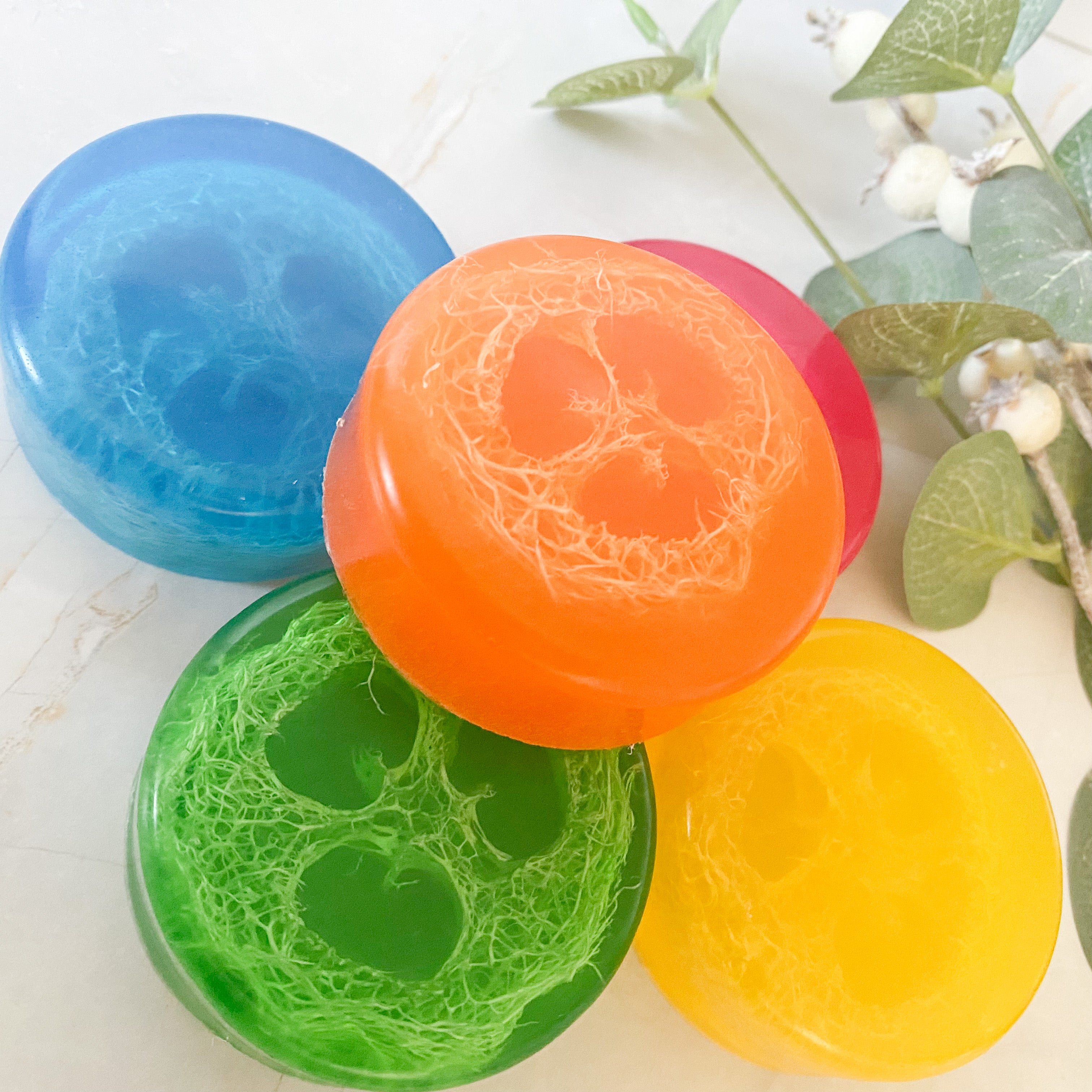 A collection of colorful loofah soaps in various colors including pink, green, yellow, orange, and blue, with natural loofah slices embedded in them.