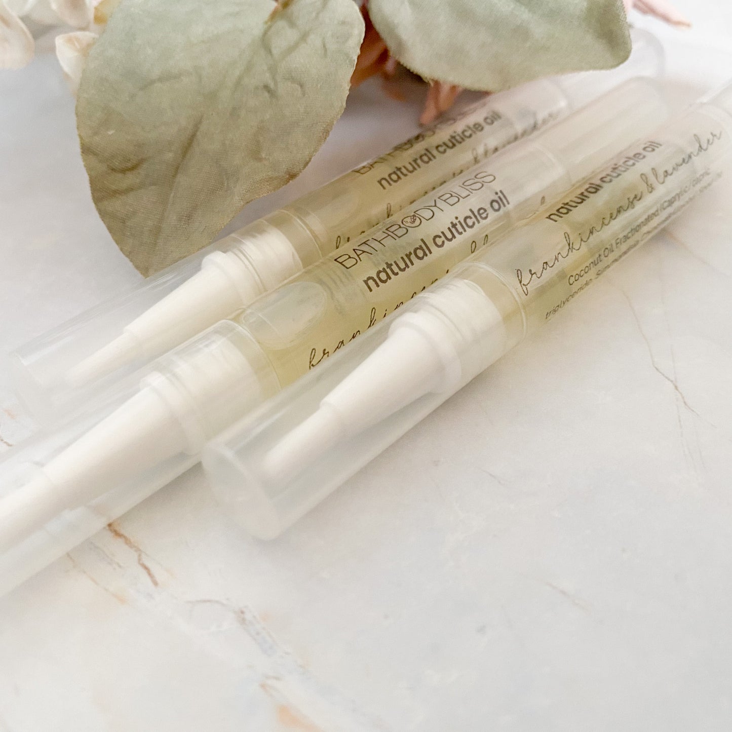 three applicator pens of natural cuticle oil on a marble surface with green leaves in the background.