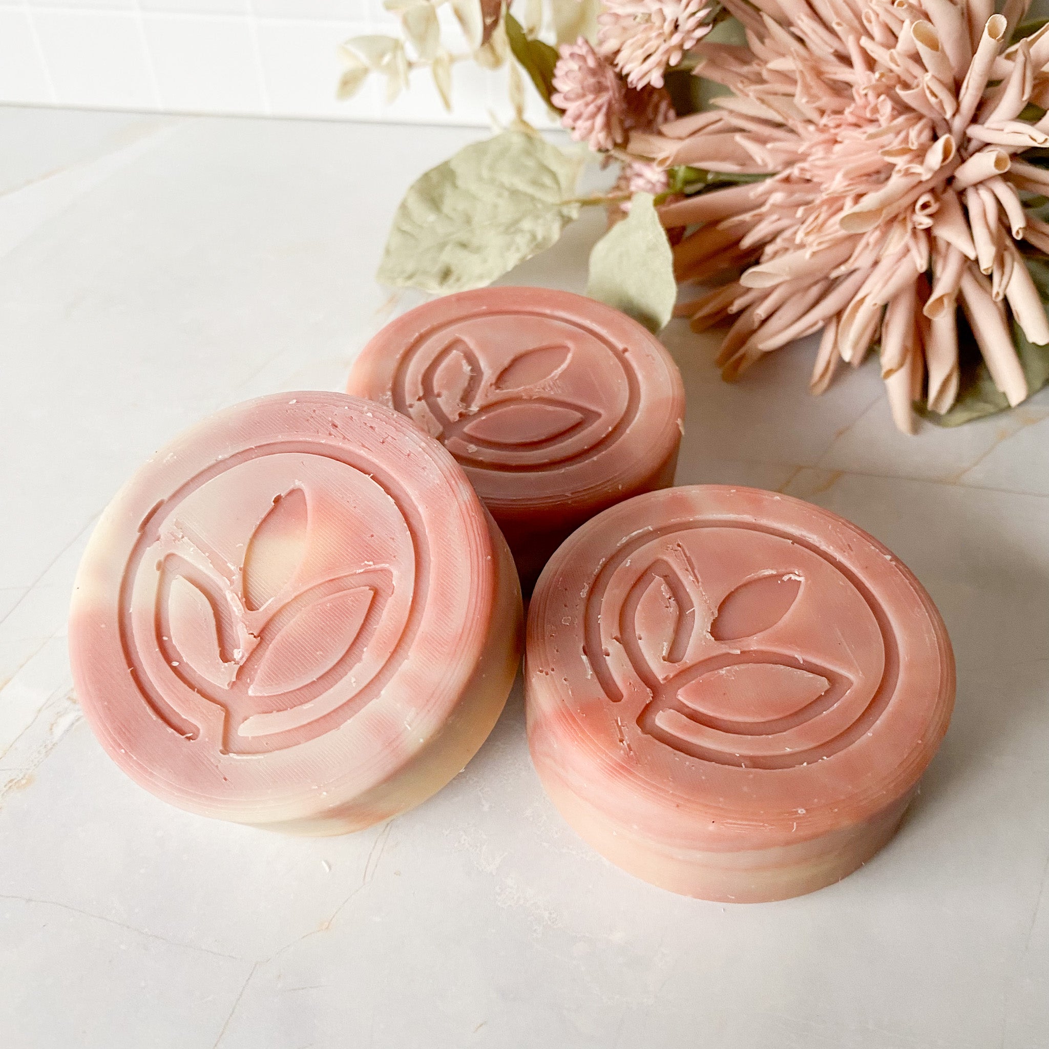 Three pink soaps with a leaf design on a white surface with flowers in the background