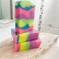 A stack of colorful handmade soap bars with a visible rainbow pattern, branded by Bathbodybliss.