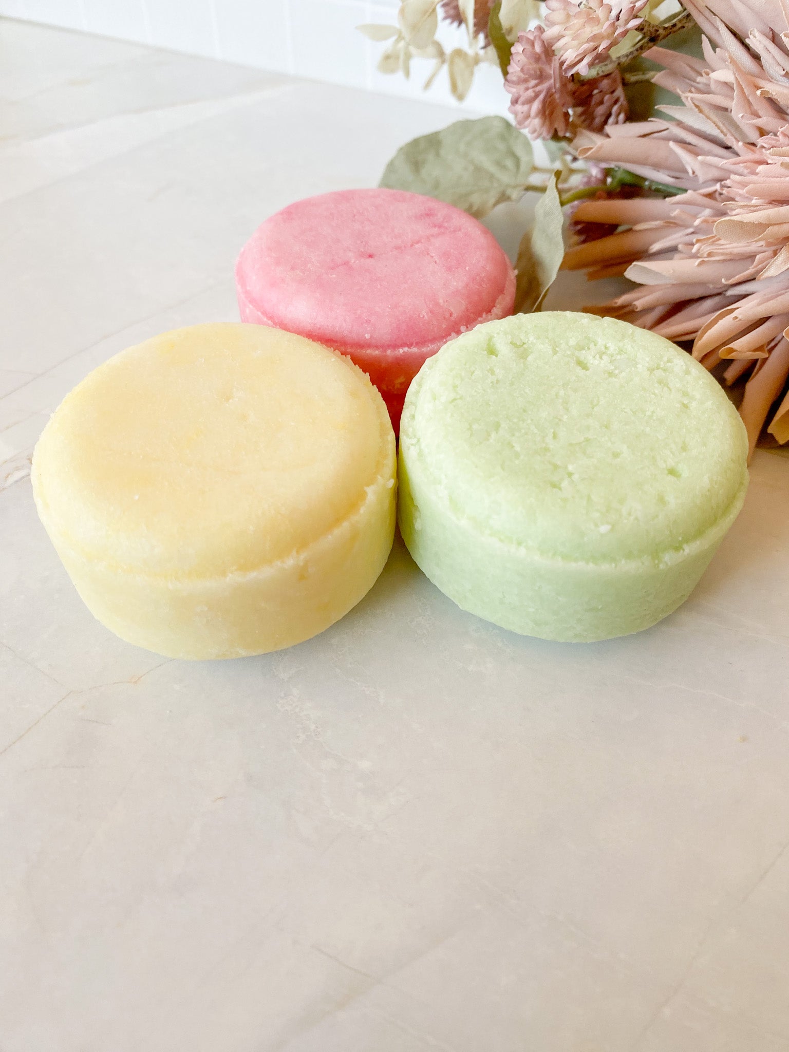 Three pastel-colored shampoo bars on a white surface with dried flowers in the background.