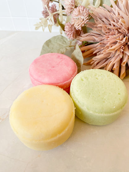 three colourful shampoo bars on a light surface with flowers in the background