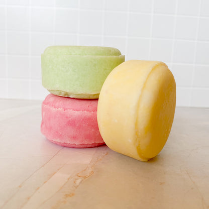 Three colorful shampoo bars stacked on a marble surface with a white tiled wall background