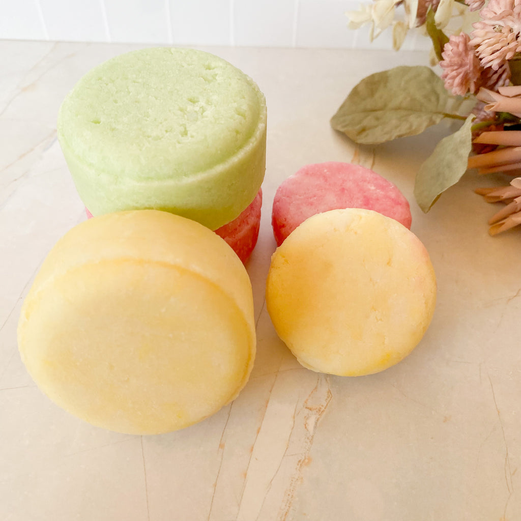 Five colourful shampoo bars on a light marble surface with a floral arrangement in the background.