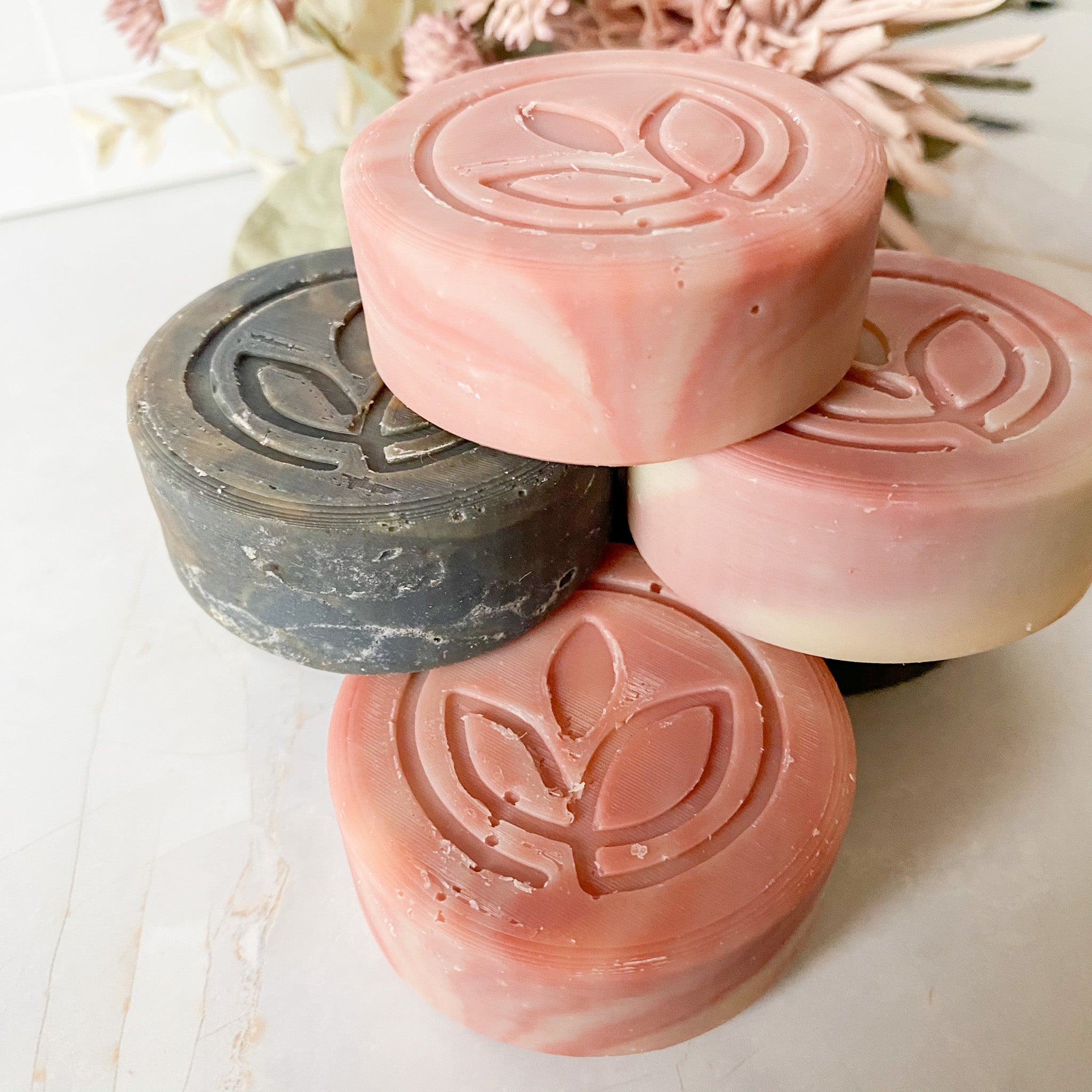 Four bars of soap with a leaf design on a light surface with flowers in the background