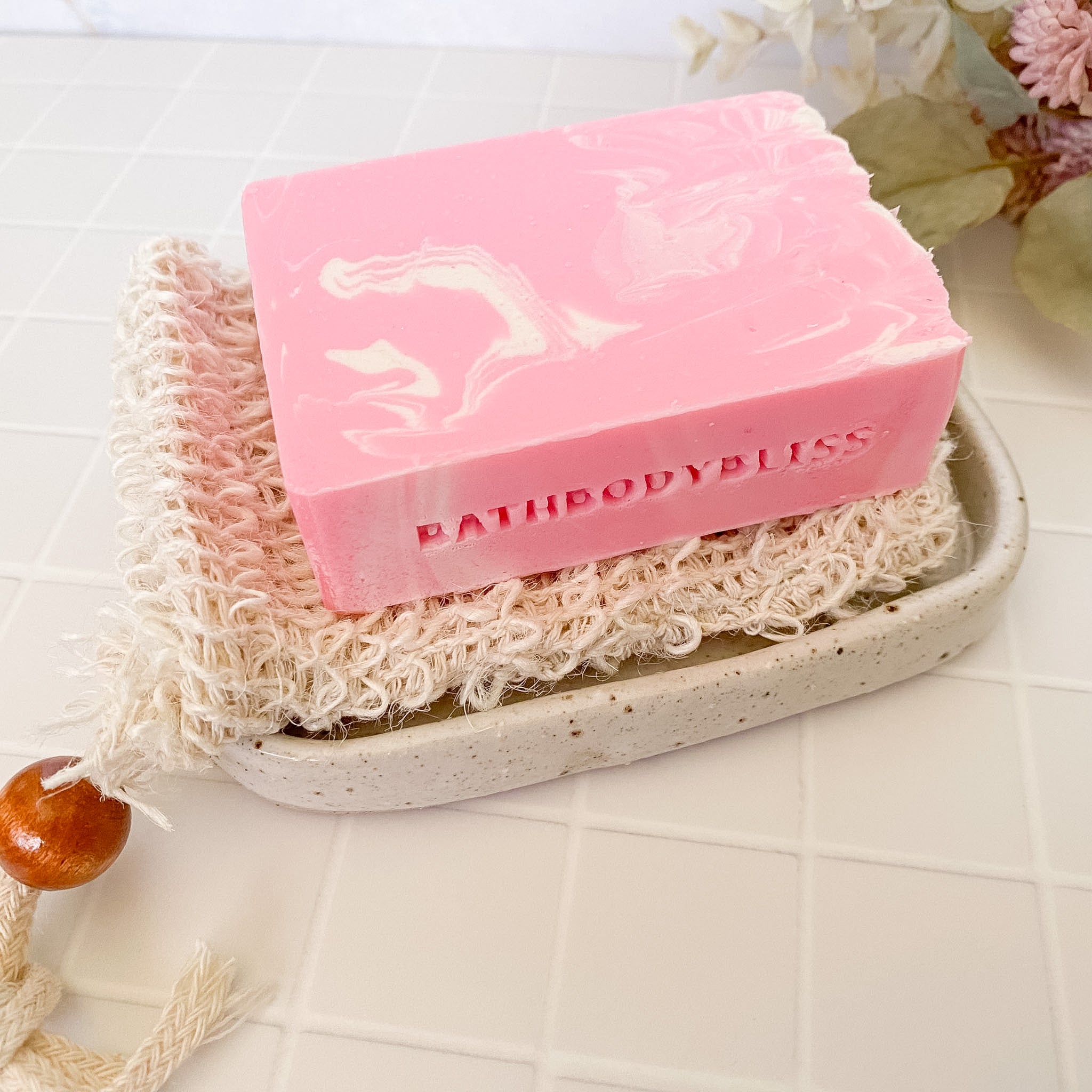 Pink soap bar with 'BATHBODYBLISS' branding on a textured soap dish with a loofah, on a light background.