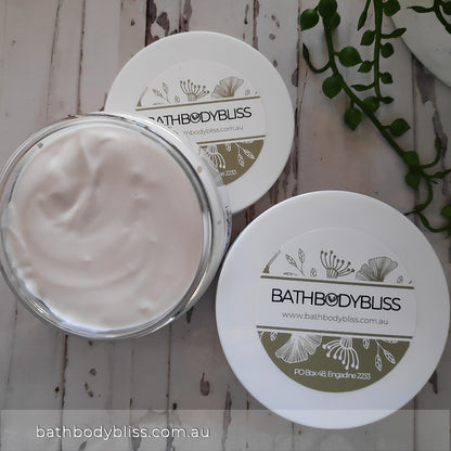 two jars of white cream with bath body bliss branded labels, on a pale wooden background with a plant in the corner