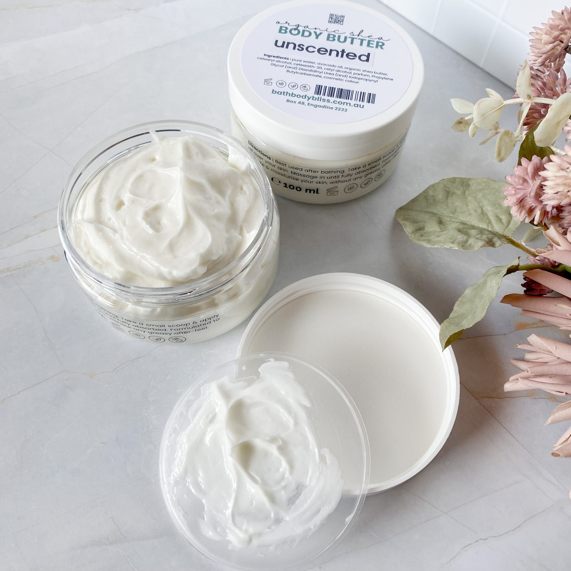 one opened jar and one closed jar of thick white body butter on a light surface with floral decor