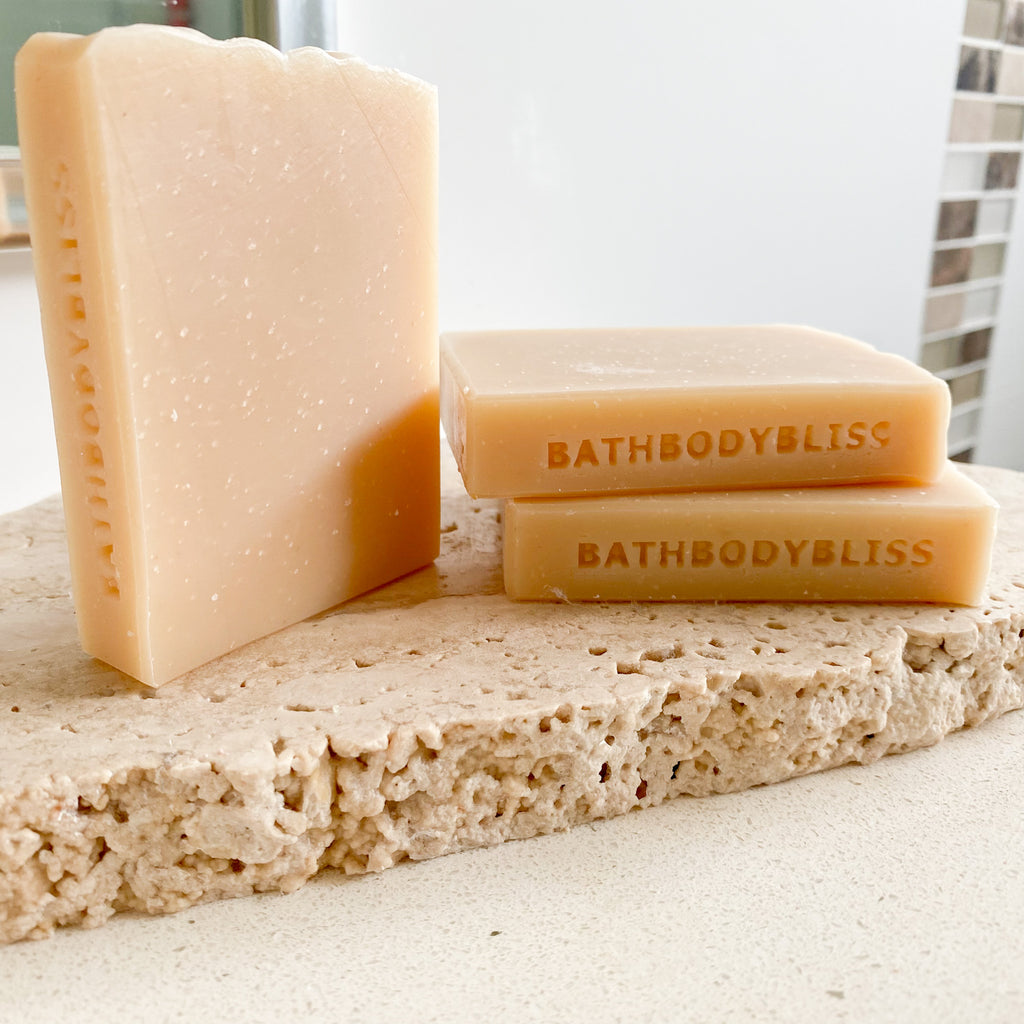 three bars of natural coloured soap, stamped with bathbodybliss logo, arranged on a rustic stone with tiled bathroom in the background