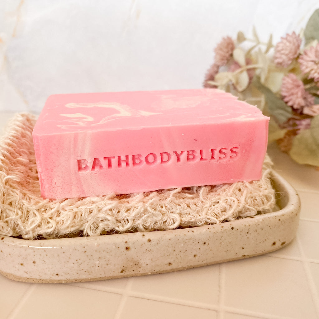 Pink soap bar with 'BathBodyBliss' text on a natural soap dish, with flowers in the background.