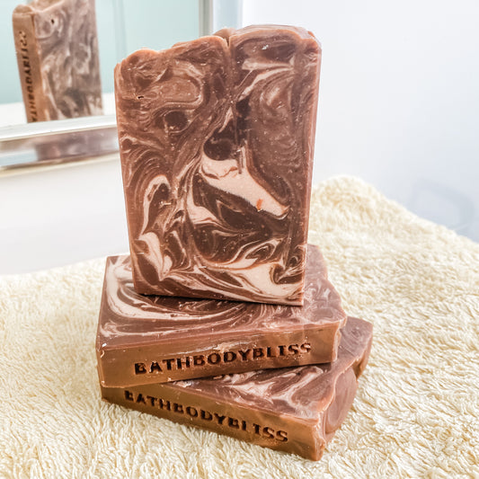 Three swirled brown and white soap bars on a textured surface with 'BathBodyBliss' branding.