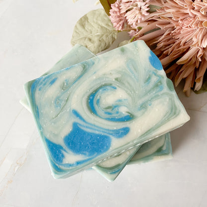 Swirled blue and green soap bars on a white surface with pink flowers in the background