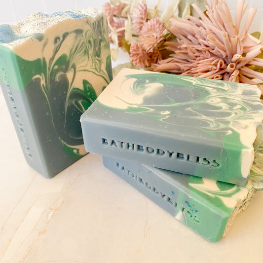 Three bars of soap with green, blue and white swirls, branded 'BathBodyBliss', on a light surface with flowers in the background.